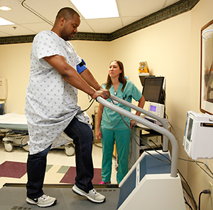 Healthcare provider performing cardiac stress test on man walking on treadmill in exam room.